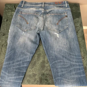 Blå slitna jeans från Dondup, stl w32 och L30 - Säljer ett par blå jeans från Dondup i storlek 32. Jeansen har en straight passform och är rejält slitna med flera hål och slitningar framtill för en cool, avslappnad look. Klassisk femficksmodell med orangea sömmar och detaljer.