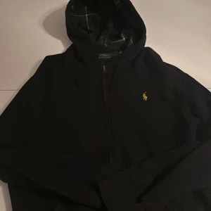 Svart hoodie från Polo Ralph Lauren M - Svart hoodie från Polo Ralph Lauren med dragkedja och huva. Insidan av huvan har rutigt foder i grönt, gult och rött. Klassisk gul broderad logga på bröstet. Perfekt för en chill och stilren look. Storlek M