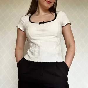T-shirt med svart kant och rosett - Supersöt vit ribbad topp med korta ärmar, svart kant runt den fyrkantiga urringningen och en liten svart rosett framtill. Figurnära passform🥰
