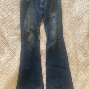 Bootcut jeans från Boosley, blå - Säljer ett par snygga blå bootcut jeans från Boosley med fyra fickor och kontrastsömmar. Jeansen har en mörk tvätt med lätt slitning framtill och bak, samt metallknapp med Boosley-logga. Perfekt för dig som gillar retrovibbar och bredare benslut.