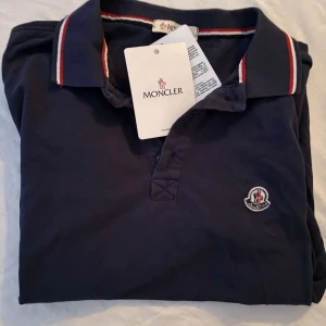 Mörkblå Moncler pikétröja - Snygg mörkblå pikétröja från Moncler med klassisk krage och korta ärmar. Tröjan har Moncler-logga på bröstet och kragen har vita och röda ränder som ger en sportig vibe. Tillverkad i mjuk bomull som är skön mot huden.                                                                                                                                                                              Nypris: 3600kr GORILLAPRIZE:950kr            Skick:7/10 rheabiliserad         KVITTO MEDKOMMER     