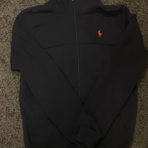 Svart hoodie från Polo Ralph Lauren - Svart hoodie från Polo Ralph Lauren med röd broderad logga på bröstet. Tröjan har dragkedja framtill, långa ärmar och ribbade muddar. Tillverkad i mjuk bomull som är skön att ha på sig. Perfekt för dig som gillar stilren streetwear.