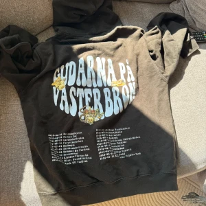 hov1 merch - Svart hoodie med stor vit tryckt text 'Gudarna på Västerbron' och turnédatum på ryggen. Framsidan har en liten broderad krona 