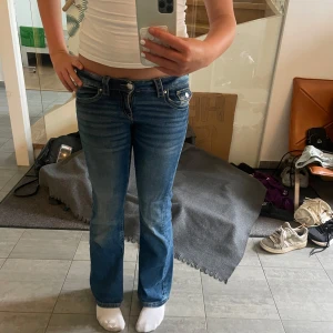 Blå bootcut jeans med låg midja - Säljer ett par blå bootcut jeans med låg midja och snygga detaljerade bakfickor med knappar. Jeansen har klassisk femficksdesign och är tillverkade i stretchigt denim för en bekväm passform. 
