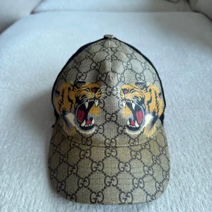 Gucci keps med tigertryck och GG-mönster - ÄKTA Snygg keps från Gucci med klassiskt GG-mönster i beige och brunt, prydd med två ilskna tigrar framtill. Baksidan har svart mesh och justerbart band i grönt och rött. Materialmix av bomull och polyester, perfekt för dig som vill sticka ut. Billigaste på StockX kostar 5000kr. 