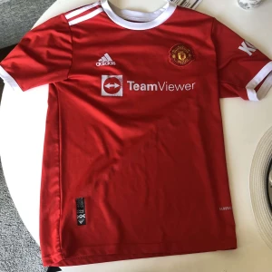 Manchester United Ronaldo 7 tröja - Säljer en röd Manchester United fotbollströja , Adidas med vita detaljer och tryck. Tröjan har korta ärmar, klubbmärke på bröstet, TeamViewer sponsortryck och Ronaldo 7 på ryggen. Materialet är lätt och andas, perfekt för match eller träning. Nummer 7 är lite bortslitet