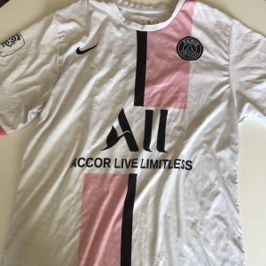 PSG Messi 30 vit fotbollströja Nike - Snygg PSG fotbollströja med Messi 30 på ryggen. Vit med svarta och rosa detaljer, PSG-logga på bröstet och Nike Swoosh. Kortärmad och tillverkad i lätt, ventilerande material. Framsidan har tryck med Accor Live Limitless och coola grafiska block i rosa och svart. På baksidan har färgen blivit lite slitet