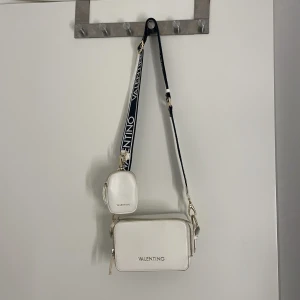 Vit axelväska från Valentino - Snygg vit axelväska från Valentino med bred svart axelrem med logga. Väskan har två fack med dragkedja och en liten matchande miniväska framtill. Guldiga detaljer och stilren design, perfekt för att lyfta din outfit. Lite missfärgad på sidorna av metallen.