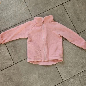 Rosa fleecejacka från Kaxs 86/92 - Mysig rosa fleecejacka från Kaxs i storlek 86/92. Jackan har dragkedja framtill, hög krage och två fickor. Perfekt för kyliga dagar och enkel att slänga på sig. Mjuk och skön känsla med långärmad passform. Hanken har lossnat i ema sidan men går lätt att sy i.