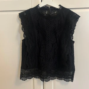 Svart spetsblus med volangdetaljer - Supersnygg svart blus i spets med vackra volangdetaljer längs ärmhål och nederkant. Hög halsringning och fina broderade mönster ger en elegant look. Perfekt för dig som gillar detaljer och vill sticka ut lite extra.