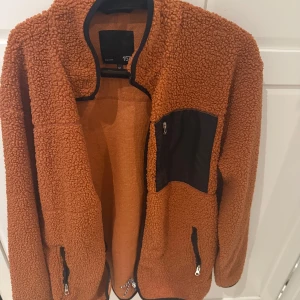 Brun teddyjacka med svarta detaljer - Mysig brun teddyjacka från 157 med svarta detaljer och dragkedjor. Jackan har en hög krage, svart bröstficka med dragkedja och två sidofickor med dragkedja. Perfekt för kyliga dagar och ger en avslappnad vibe.