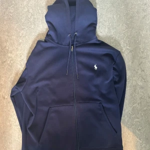 Mörkblå hoodie från Ralph Lauren - Snygg mörkblå hoodie från Ralph Lauren med dragkedja och klassisk broderad logga på bröstet. Tröjan har huva med snörning och stora fickor framtill. Perfekt för dig som gillar stilren och sportig look.