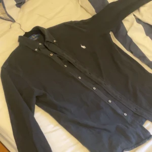 Svart skjorta från Ralph Lauren - Klassisk svart skjorta från Ralph Lauren med vit broderad logga på bröstet. Skjortan har lång ärm, knappar hela vägen och en snygg krage. Perfekt för dig som gillar stilrena och tidlösa plagg.