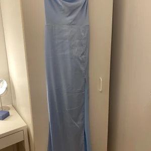 Ljusblå långklänning med slits - Supersnygg ljusblå långklänning i satinliknande material med tunna axelband som korsas i ryggen. Klänningen har en slits på sidan och öppen rygg för en extra elegant look. Perfekt för bal eller festliga tillfällen.