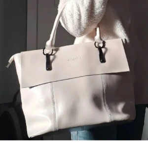 Beige axelväska från Adax - Snygg beige axelväska från Chloé i slätt skinn med stilrena metalldetaljer. Väskan har ett stort lock, dubbla handtag och en praktisk dragkedja på baksidan. Perfekt för dig som vill ha en klassisk och rymlig väska till vardagen.