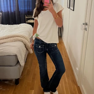 Mörkblå bootcut jeans med fickdetaljer - Säljer ett par mörkblå jeans med bootcut passform och markerade vita sömmar. Jeansen har två framfickor med lock och knapp, samt klassiska bakfickor. Perfekta för en avslappnad och trendig look.💞