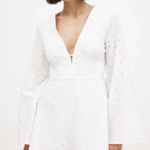 Vit klänning med öppen rygg från NA-KD - Superfin vit klänning från NA-KD med broderat blommönster, djup v-ringning och lång ärm. Klänningen har en snygg öppen rygg och markerad midja. Perfekt till sommaren och student 