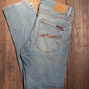 Ljusblå jeans från Nudie Jeans - Säljer ett par klassiska ljusblå jeans från Nudie Jeans med orange sömmar och signaturbroderi på bakfickan. Jeansen har normal passform, fem fickor och knappgylf. Perfekta för en avslappnad stil.