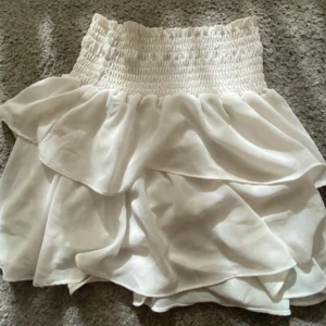 Vit volangkjol med smock i midjan - Supersöt vit kjol med volanger och smockad midja. Kjolen är kort och har en luftig känsla, perfekt för varma dagar. Materialet känns mjukt och lätt mot huden. Den har en liten fläck som kanppt syns!💕💕