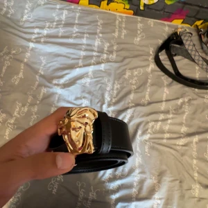 Svart bälte med guldspänne - Säljer ett svart bälte med ett stort, detaljerat guldspänne som har ett ansikte i relief. Bältet är stilrent och gjort i slätt material, perfekt för att lyfta en outfit.