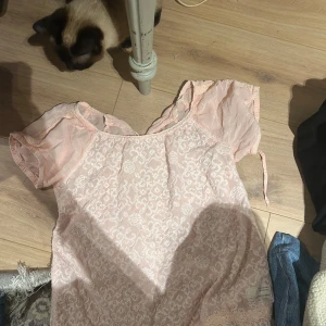 Ljusrosa blus med spetsdetaljer - Söt ljusrosa blus med korta puffärmar och spetsmönster framtill. Blusen har en romantisk känsla med volangliknande ärmar och rund halsringning. Perfekt för en feminin och somrig stil.