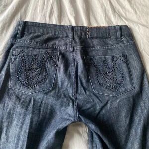 Lågmidjade mörkblå jeans med skit snygga detaljer på bakfickorna - Säljer ett par lågmidjadejeans  mörkblå jeans med bootcut och snygga strassdetaljer på bakfickorna. Klassisk femficksmodell med normal passform och knappgylf. Perfekta för dig som vill ha något extra till vardagslooken. Midjemått 36 cm rakt över. Innerbenslängd 85 cm. Lite slitna längst ned vid kanten vilket jeans ofta blir kan skicka bild privat. 