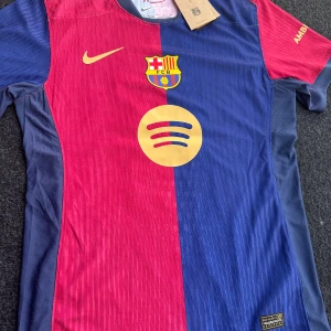 FC Barcelona matchtröja Raphinha 11 - Säljer en officiell FC Barcelona matchtröja från Nike med Raphinha och nummer 11 på ryggen. Tröjan är kortärmad, har klassiska blå och röda vertikala fält, klubbmärke på bröstet och UNHCR-logga på ryggen. Perfekt för dig som älskar Barca!