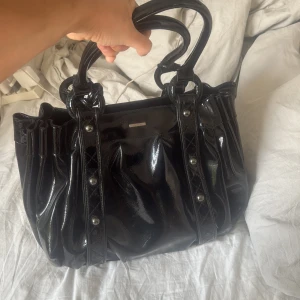 Svart glansig handväska med nitar - Säljer en svart glansig handväska med dubbla handtag och dekorativa nitar längs sidorna. Väskan har flera innerfack och rymmer det mesta du behöver till vardagen. Perfekt accessoar för dig som gillar en stilren men edgy look.
