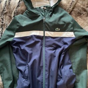 Grön och blå vindjacka från Lacoste - Säljer en snygg vindjacka från Lacoste i grönt och blått med vit detalj och dragkedja. Tunn jacka vilket är skönt nu när det börjar bli lite varmt, hör av dig vid intresse eller frågor 😁