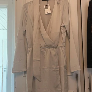 Beige omlottklänning från NA-KD - Säljer en beige omlottklänning från NA-KD med lång ärm och knytband i midjan. Klänningen har v-ringning och är tillverkad i ett mjukt, slätt material. Perfekt för dig som gillar stilrena och enkla plagg.