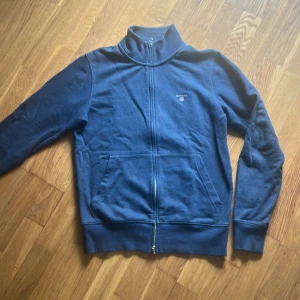 Marinblå ziptröja från GANT - Säljer en marinblå ziptröja från GANT med hög krage och dragkedja hela vägen. Tröjan har två fickor framtill och diskret GANT-logga broderad på bröstet. Perfekt för lager-på-lager och snygg till jeans eller chinos.