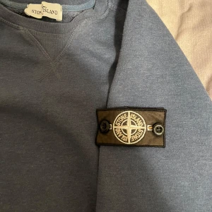 Blå sweatshirt från Stone Island - Säljer en blå sweatshirt från Stone Island med klassisk logotyp-patch på vänster ärm. Tröjan har rund halsringning och långa ärmar. Perfekt för en avslappnad och stilren look. Passar även M