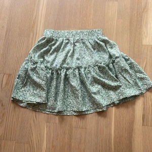 Grön blommig kjol från SHEIN - Söt grön kjol från SHEIN med vitt blommönster och volangdetalj. Kjolen har en kort längd och resår i midjan för en bekväm passform. Perfekt för dig som gillar romantiska och somriga plagg.