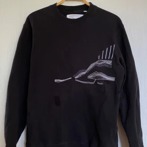 Svart sweatshirt från L'Homme Rouge med broderad detalj - Svart sweatshirt från L'Homme Rouge med unik vit broderad design på framsidan. Tröjan har rund halsringning och långa ärmar. Perfekt för dig som gillar stilrena plagg med en twist. OBS. Den svarta ”fläcken” är bara vatten. Du kan vara lugn 