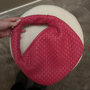 Flätad rosa axelväska - Säljer en snygg axelväska i starkt rosa färg med flätad design. Väskan har en rundad form och ett kort handtag, perfekt för att bära över axeln men även i handen. Och defensivt till en fin balklänning. Stängs med dragkedja och är tillverkad i syntetmaterial. Nypris 600kr. 