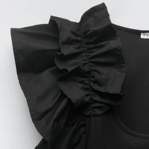 Svart croppad volangtopp med ärmlös design - Snygg kort svart topp med stora volanger längs axeln och ärmlös design. Rund halsringning och mjukt material.