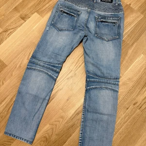 Ljusblå bikerjeans från Balmain size 32  - Säljer ett par ljusblå bikerjeans från Balmain med dragkedjor på fickorna och ribbade detaljer vid knäna och höften. Jeansen har raka ben och klassisk femficksdesign. Perfekta för dig som vill sticka ut med en cool streetstil.