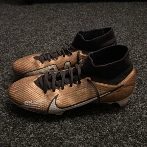 Guldiga Nike fotbollsskor med högt skaft - Säljer ett par guldiga Nike fotbollsskor med svart högt skaft och svarta skosnören. Skorna har en vit Nike-logga på sidan och dobbar undertill för bra grepp på både konstgräsplan och gräsplan. Pris kan diskuteras.