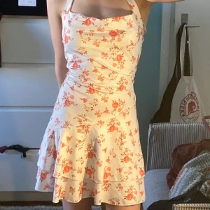 Vit blommig kortklänning med volang - Supersöt vit klänning med rosa blommigt mönster och volangdetaljer nertill. Axelbandslös modell med figurnära passform upptill och utsvängd kjol. Perfekt för sommarens alla tillfällen!