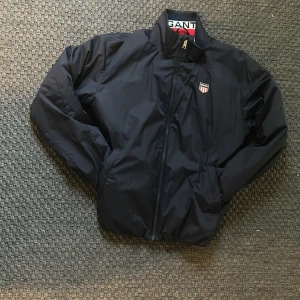 Svart vindjacka från GANT - Snygg svart vindjacka från GANT med dragkedja och ståkrage. Jackan har GANT-logga på bröstet och elastiska muddar vid ärmslut och midja. Perfekt för blåsiga dagar och enkel att matcha med olika outfits. Storlek 176/S men passar M