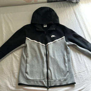 Nike hoodie jacka med dragkedja - Snygg hoodie jacka från Nike i svart och grått med hel dragkedja och huva. Jackan har vita detaljer, två fickor fram och en extra ficka med dragkedja på ärmen. Perfekt för en sportig look.