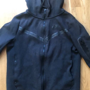 Svart hoodiejacka från Nike - Svart hoodiejacka från Nike med dragkedja framtill och diskreta svarta detaljer över bröstet. Jackan har huva, lång ärm och två fickor framtill samt en extra ficka med dragkedja på ärmen. Perfekt för en sportig look.