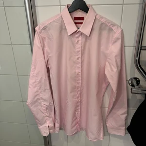 Ljusrosa skjorta från Hugo - Säljer en stilren ljusrosa skjorta från Hugo med klassisk krage och knäppning framtill. Skjortan har lång ärm och är perfekt för dig som vill ha en clean och snygg look. Passar till många olika outfits! Denna är lite ful i färgen vid kragen, kanske går att lösa i tvätten - därför jag säljer billigt. Passar Small/medium.