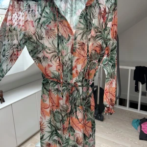 Blommig långklänning från Pernilla Wahlgren - Säljer en luftig och transparent långklänning från Pernilla Wahlgren i storlek XS. Klänningen har blommigt mönster i grönt, orange och rosa, knyt i midjan och långa ärmar. Perfekt för sommarens alla tillfällen!