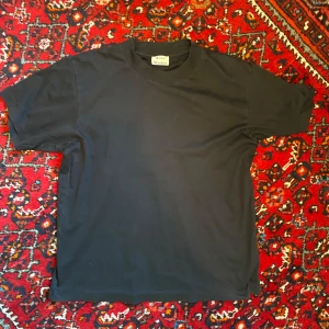 Svart t-shirt från Acne Studios - En klassisk svart t-shirt från Acne Studios i storlek L. T-shirten är tillverkad i mjuk bomull och har en enkel, rak passform med rund halsringning och korta ärmar. Perfekt basplagg för garderoben. Har ett pyttelitet hål på framsidan (sista bilden), annars bra skick.