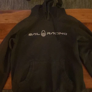 Svart hoodie från Sail Racing - Svart hoodie från Sail Racing med vit logga på bröstet. Klassisk modell med huva och känguruficka framtill. Perfekt för en avslappnad stil och enkel att matcha med det mesta.