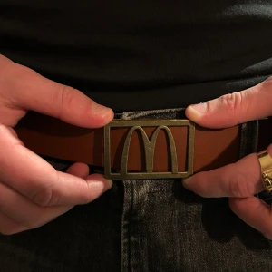 McDonald’s bälte - Säljer ett brunt bälte med ett stort metallspänne som har en unik design. Bältet är gjort i skinn och passar perfekt till jeans eller andra byxor. Snygg detalj som sticker ut och ger outfiten en cool touch.