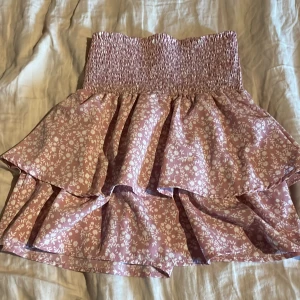 Rosa blommig volangkjol från Seaside - Supersöt rosa kjol från Seaside med vitt blommönster och bred smock i midjan. Kjolen har dubbla volanger som ger en fin och luftig känsla. Perfekt för dig som gillar romantisk stil och vill ha något bekvämt och snyggt.