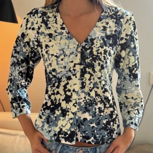 Mönstrad blus med v-ringning - Säljer en blus med abstrakt mönster i blått, svart, vitt och beige. Blusen har v-ringning, knappar framtill och trekvartsärm. Perfekt för dig som gillar unika mönster och vill sticka ut.