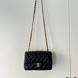 Svart quiltad axelremsväska - Säljer en klassisk svart mini axelremsväska i quiltat skinn med guldfärgade detaljer och kedjerem. Väskan är köpt utomlands och den är inte äkta. Väskan har guld spänne framtill som har tappat lite färg och röd insida med dragkedjefack. Perfekt för dig som vill ha en tidlös och lyxig accessoar. Kan användas som handväska genom att göra en ”knut” av bandet på insidan. Priset kan diskuteras. 